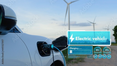 Electric car recharging energy from EV charging station display futuristic smart battery status hologram by EV charger plug cable in wind turbine farm. Alternative clean energy sustainability. Peruse © Summit Art Creations