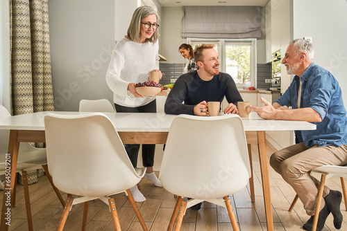 Cheerful senior woman serving table while her son talking with father