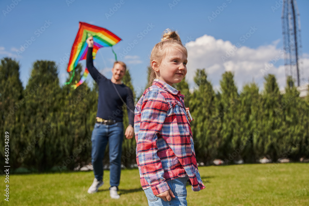 Little blonde girl standing at summer green meadow while playing with father