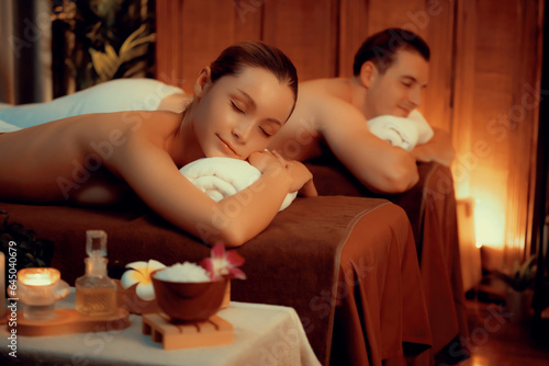 Caucasian couple customer enjoying relaxing anti-stress spa massage and pampering with beauty skin recreation leisure in warm candle lighting ambient salon spa at luxury resort or hotel. Quiescent