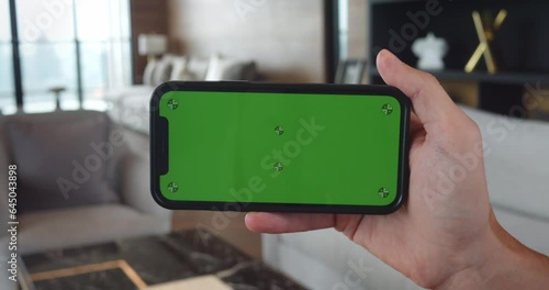 Smartphone with green chroma key mock up screen in with horizontal position at home background. Male hand hold smartphone with chroma key green screen at livingroom. Watching internet video concept