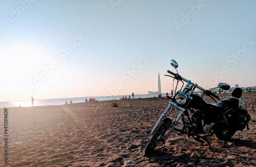 Ride by the Sea: Vintage Motorcycle at Sunset on Beach. A timeless classic motorcycle parked on the seaside at sunset, with people lounging on the beach and newly built structures in the backdrop.