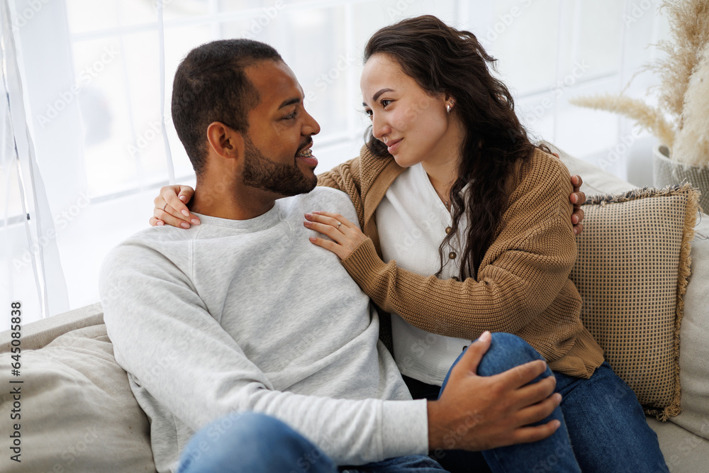 Smiling young asian woman hugging african american boyfriend while relaxing together on couch at home Smiling young asian woman hugging african american boyfriend while relaxing together on couch at home