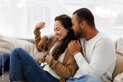 Smiling african american man hugging and talking to asian girlfriend while relaxing on couch at home © Dmytro Hai