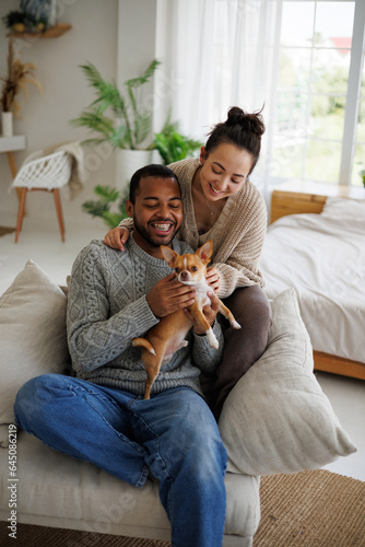 Happy african american man in sweater holding chihuahua dog near asian girlfriend on armchair at home