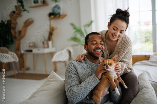Positive young multiethnic couple in sweaters petting chihuahua dog on armchair at home