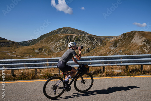 Woman cyclist is riding in the mountains and drinking water during her training with amazing view. Female cyclist in cycling kit and a helmet drinks water from a sports bottle.Transalpina, Romania