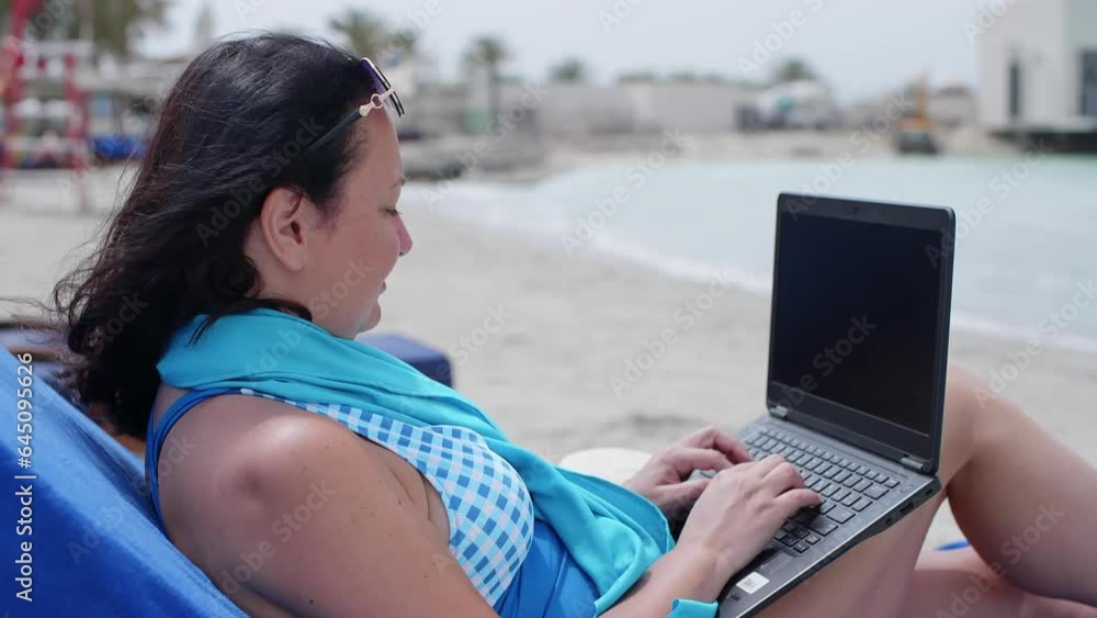 Freelancer typing on laptop while resting on lounge chair. Woman in swimwear working remotely at beach. She is relaxing on seashore during windy day in summer. Freelancer typing on laptop while resting on lounge chair. Woman in swimwear working remotely at beach. She is relaxing on seashore during windy day in summer.