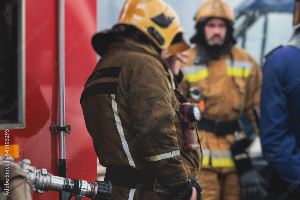 Obraz premium Group of fire men in protective uniform during fire fighting operation in the city streets, firefighters brigade with the fire engine truck vehicle in the background, emergency and rescue service