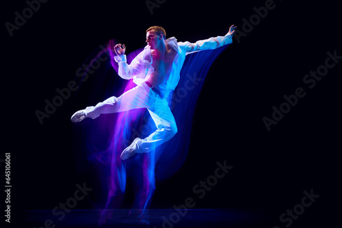 Fototapeta Naklejka Na Ścianę i Meble -  Dynamic image of stylish young man in white clothes humping over black studio background in neon with mixed lights effect. Concept of movements, art, dance and sport, fashion, youth. Copy space for ad