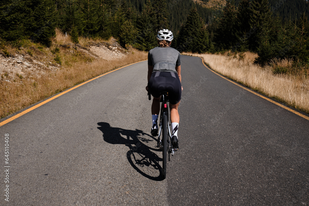 Fototapeta premium Close-up back view of a woman cyclist training on a mountain road. Cycling in the woods.Professional female cyclist is riding on a forest asphalt road.Fit athlete wearing sportswear and helmet.Romania