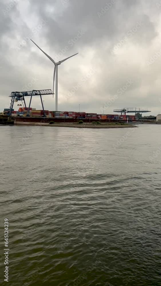 Slow motion views traveling past a wind turbine and industrial freight yard. Located on the Waal River in the Netherlands. Captured while river cruising through the Dutch countryside. Slow motion views traveling past a wind turbine and industrial freight yard. Located on the Waal River in the Netherlands. Captured while river cruising through the Dutch countryside.