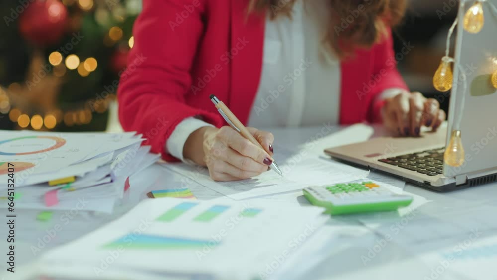 Christmas time. Closeup on middle aged business woman in santa hat and red jacket with documents and laptop working in green office with Christmas tree. Christmas time. Closeup on middle aged business woman in santa hat and red jacket with documents and laptop working in green office with Christmas tree.