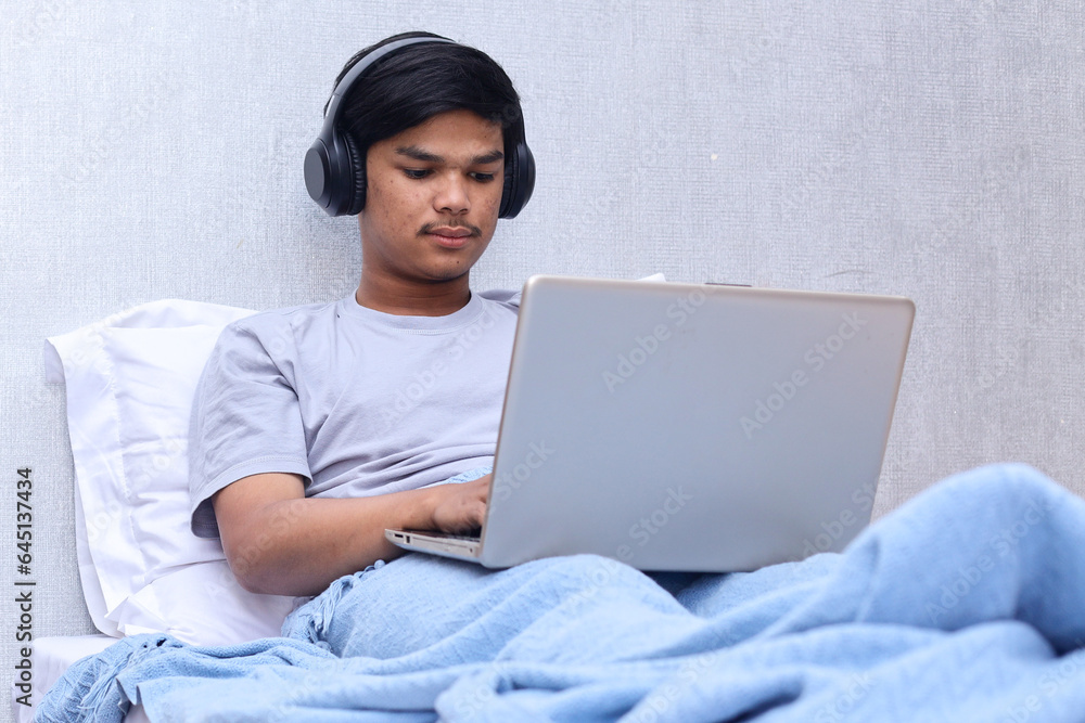 Young man wearing headphones and working on laptop while sit on bed at bedroom Young man wearing headphones and working on laptop while sit on bed at bedroom