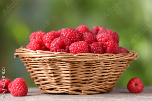Wicker basket with tasty ripe raspberries on wooden table against blurred green background, closeup