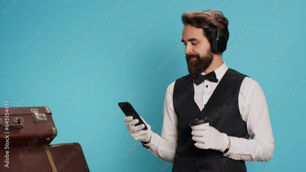 Doorman texting messages drinking coffee in studio, enjoying break with music on headphones. Young adult with classy hotel sector job messaging and sipping on cold brew beverage. Doorman texting messages drinking coffee in studio, enjoying break with music on headphones. Young adult with classy hotel sector job messaging and sipping on cold brew beverage.