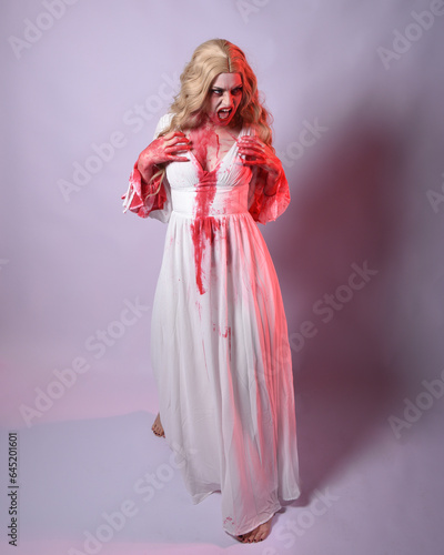  Full length portrait of scary vampire zombie bride, wearing elegant halloween fantasy costume dress with bloody red paint splatter. standing walking pose. Isolated on white studio background 
