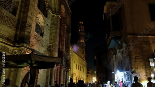 People walking around streets of Khan El Khalili bazaar in Cairo, Egypt. Night Cairo street. Minaret of Complex of Sultan al-Mansur Qalawun at background photo
