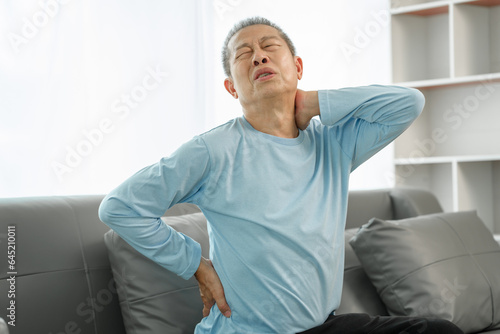 Elderly Asian man on sofa, grimacing from spinal pain, seeking relief from osteoporosis-related discomfort at home. © makibestphoto