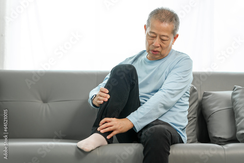 Senior Elderly Asian man in blue shirt suffering from intense foot and ankle pain on the sofa. photo
