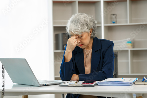 Mature businesswoman at her desk, overwhelmed by stress, feeling the weight of failure, and suffering from a headache, Stressful workday, Overwhelmed at work, Corporate crisis