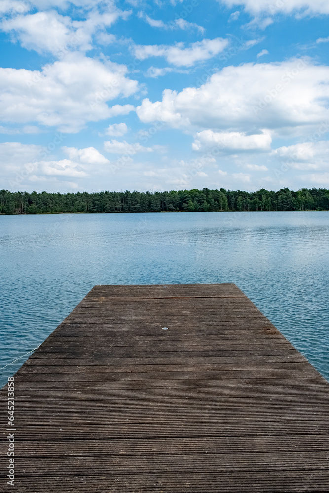 A serene wooden pier stretches out over the pristine waters of a tranquil forest lake, set against a backdrop of fluffy white clouds in a pristine sky. This idyllic scene invites you to escape the