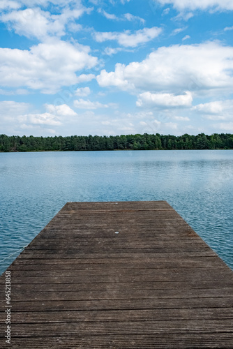 A serene wooden pier stretches out over the pristine waters of a tranquil forest lake, set against a backdrop of fluffy white clouds in a pristine sky. This idyllic scene invites you to escape the