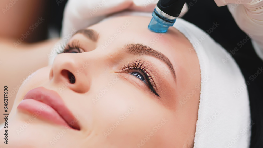 A beautiful, young woman undergoes a vacuum hydro-peeling procedure in a beauty salon on a light background.