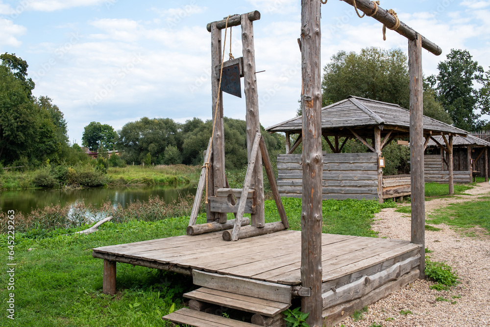 A guillotine on a wooden platform, behind it a river and a wooden barn. A guillotine on a wooden platform, behind it a river and a wooden barn.