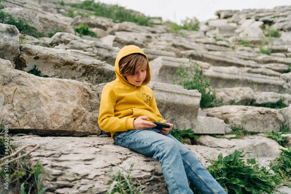 Fototapeta premium School kid child playing with his cellphone smartphone in an ancient stone amphitheater. Boy in yellow hoodie texting over his phone gadget while sitting on a stone bench in abandoned antique arena.