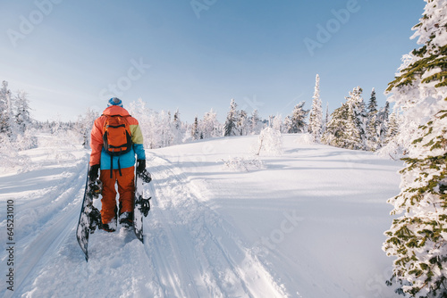 Male snowboarder wearing orange clothing walking in snow covered spruce forest on sunny day, holding two snowboards