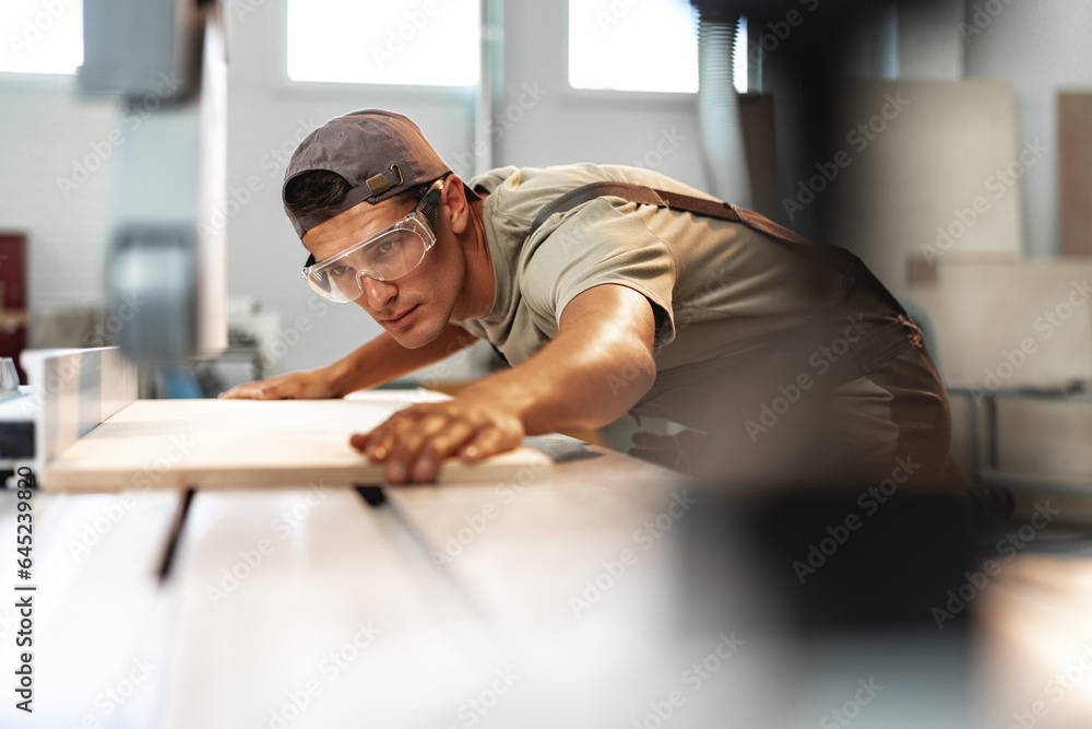 © fotofabrika - Young carpenter cutting a piece of wood in using a circular saw in furniture factory