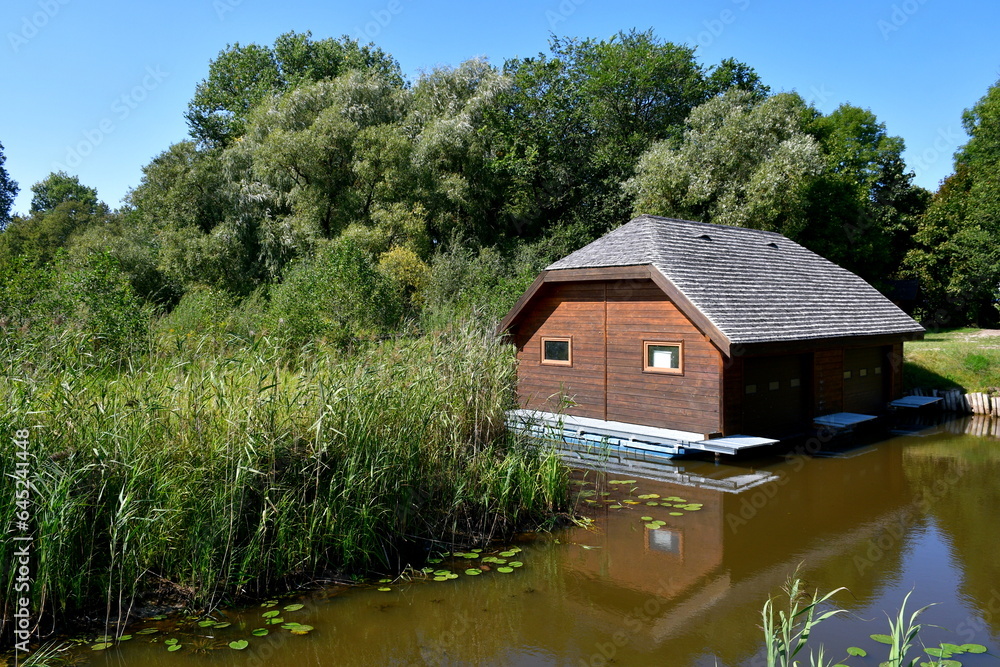 A view of a small house, shack, or shelter with an angled roof made out of thatch located next to a shallow pond or river overgrown with reeds, grass, and other flora seen in Poland in summer A view of a small house, shack, or shelter with an angled roof made out of thatch located next to a shallow pond or river overgrown with reeds, grass, and other flora seen in Poland in summer