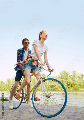 Vertical portrait of active young couple riding one bicycle on city street. Lovely, nice riders enjoying themselves. Romantic leisure activity.