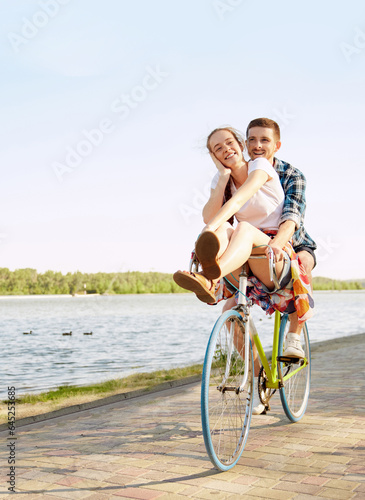 Vertical portrait of active young couple rejoicing riding one bicycle on embankment. Lovely, nice cyclists in love enjoying walk. Romantic leisure activity.