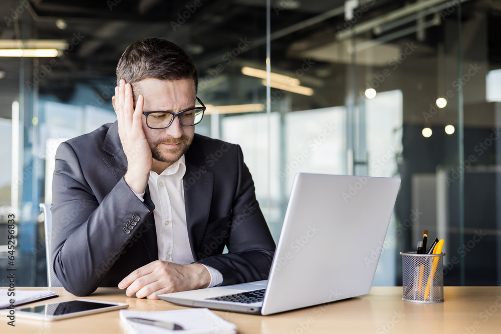 Problems at work. Upset young male office worker, businessman sitting at a desk in front of a laptop and thoughtfully leaning his head on his hand Problems at work. Upset young male office worker, businessman sitting at a desk in front of a laptop and thoughtfully leaning his head on his hand