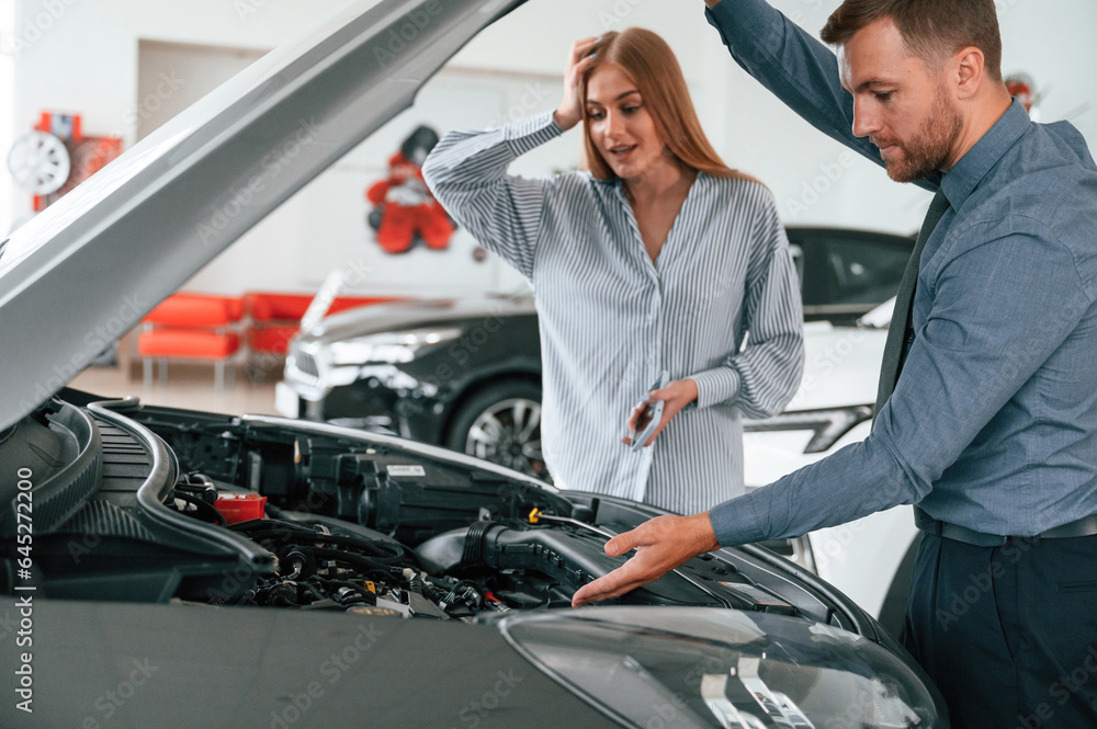 Under the hood. Man in formal clothes is with woman customer with the electric car Under the hood. Man in formal clothes is with woman customer with the electric car
