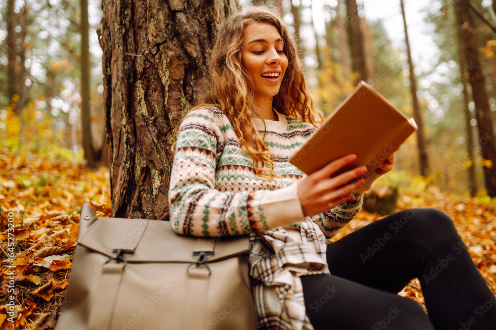 Obraz premium Curly-haired woman in a stylish sweater with a book sits under a maple tree in the autumn forest, reads, enjoys the weather. Concept of autumn, holiday.