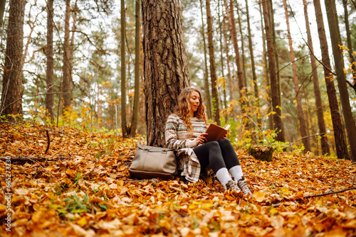 Curly-haired woman in a stylish sweater with a book sits under a maple tree in the autumn forest, reads, enjoys the weather. Concept of autumn, holiday. © maxbelchenko