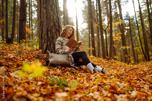 Curly-haired woman in a stylish sweater with a book sits under a maple tree in the autumn forest, reads, enjoys the weather. Concept of autumn, holiday.