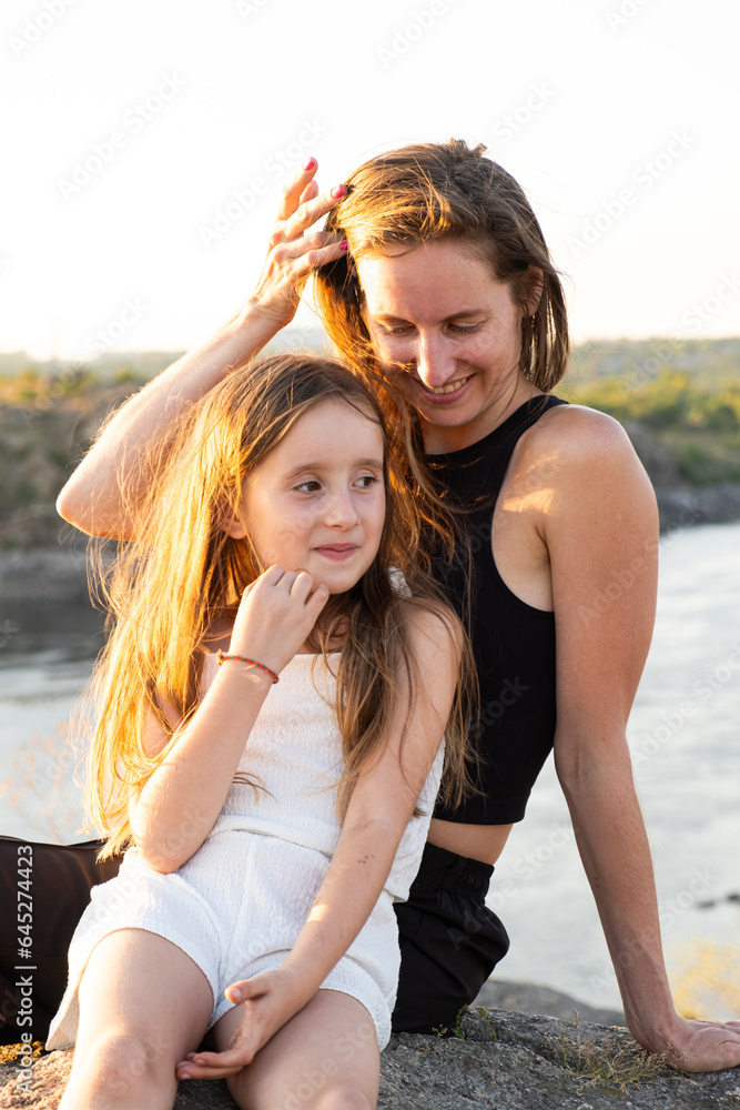 Happy mom and daughter relaxing on rocks in nature against the backdrop of the river. Happy Motherhood, Mother's Day Happy mom and daughter relaxing on rocks in nature against the backdrop of the river. Happy Motherhood, Mother's Day