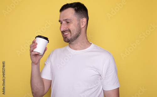 Young caucasian smiling bearded attractive man drinking hold paper cup of coffee isolated on yellow background studio portrait. People lifestyle concept.