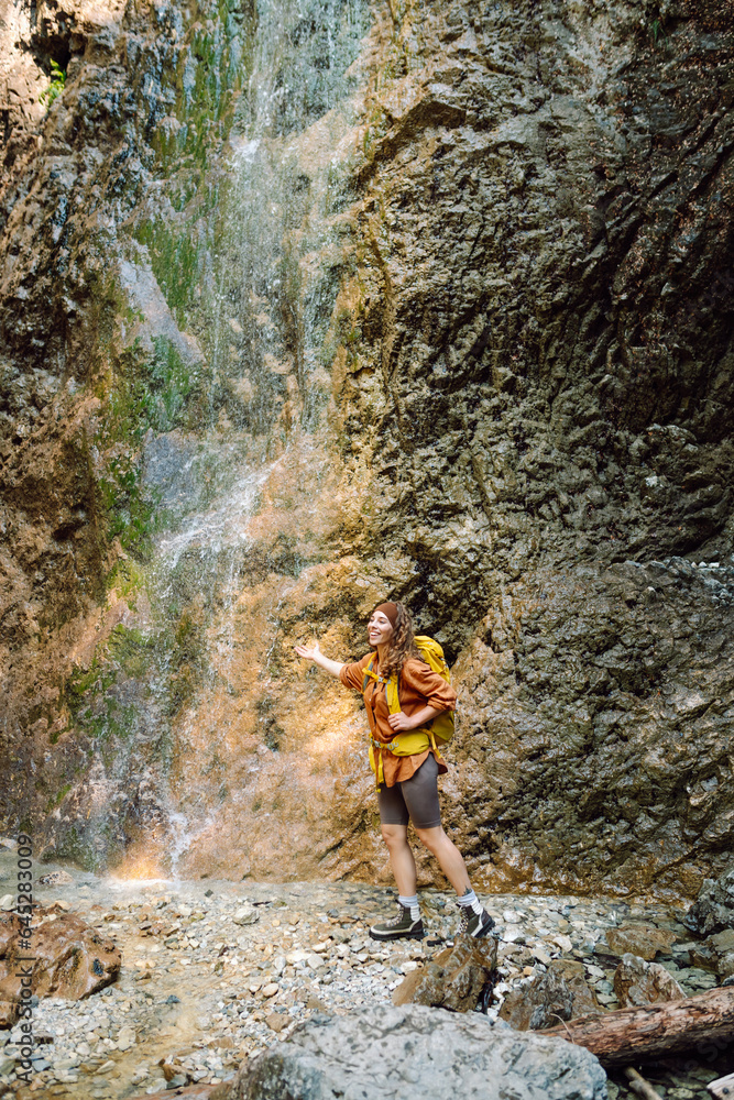 Smiling female traveler with a yellow backpack, dressed in hiking clothes, enjoys a waterfall near a mountain river. Travel, trekking. Nature concept.