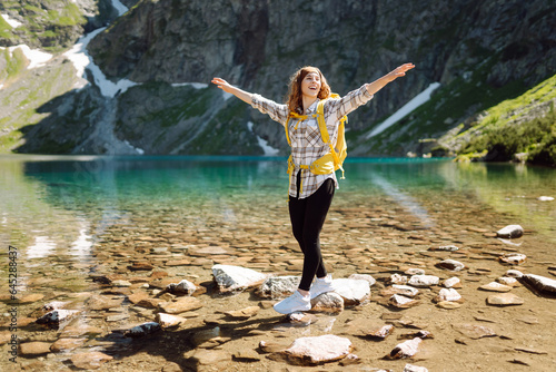A smiling female traveler with a yellow hiking backpack against the backdrop of a turquoise lake among the mountains. Active lifestyle. Hiking  adventure concept.