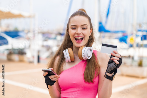 Young pretty sport girl at outdoors buying with the mobile and holding a credit card with surprised expression