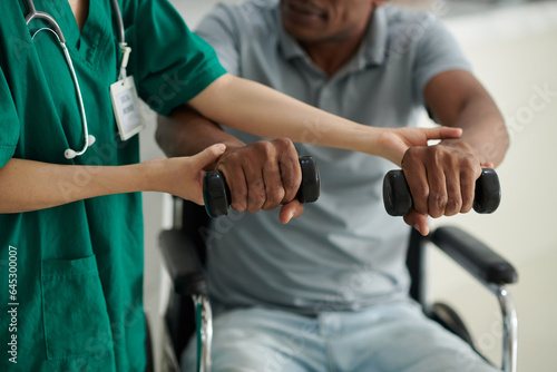 Closeup image of man with disability exercising with dumbbells under control of social worker