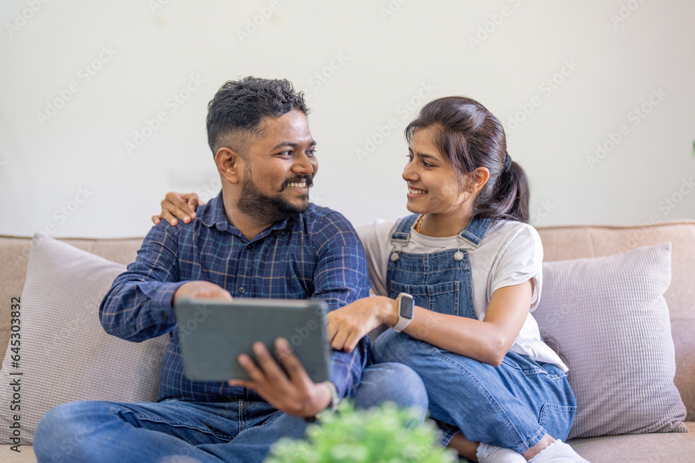 Beautiful indian spouses rest on sofa at home, using digital tablet explore the world of social media and shopping online together