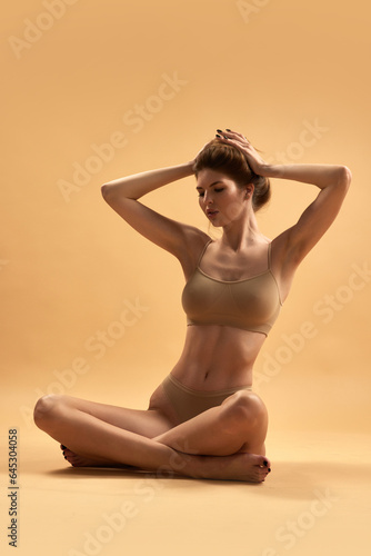 Portrait of young girl with slim body sitting in lotus pose in beige inner wear and looking at camera against sand studio background