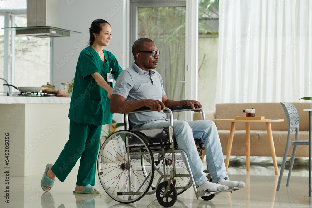 Smiling young social worker pushing wheelchair of patient with disability