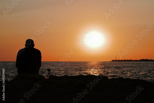 Signore che guarda e filma il tramonto da una scogliera a Marina di Ragusa photo